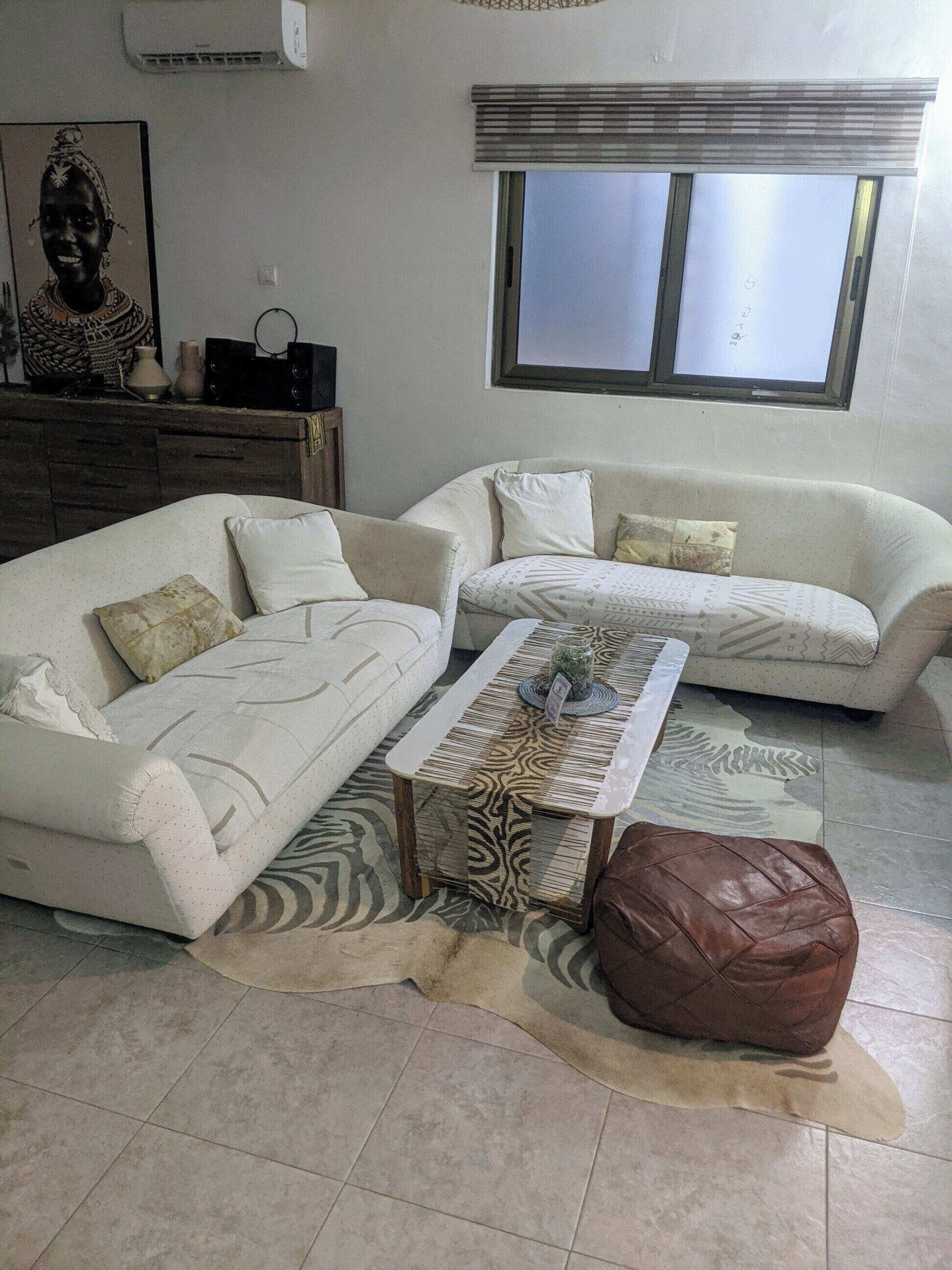 A living room with two sofas, a coffee table, a brown ottoman, a rug, and a window with blinds.