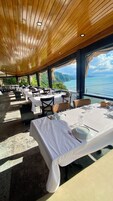 An indoor dining area with tables set for service, large windows offering a view of the sea and hills, and a wooden ceiling.