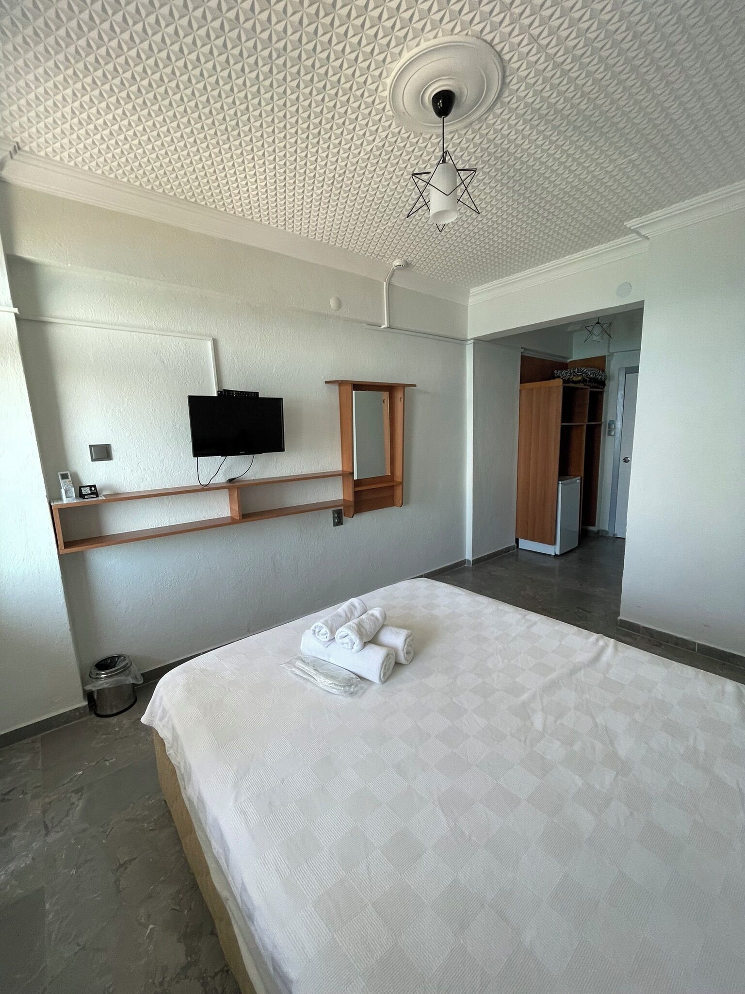 A hotel room with a bed, a TV mounted on the wall, a wooden shelf, a mirror, and a wardrobe.