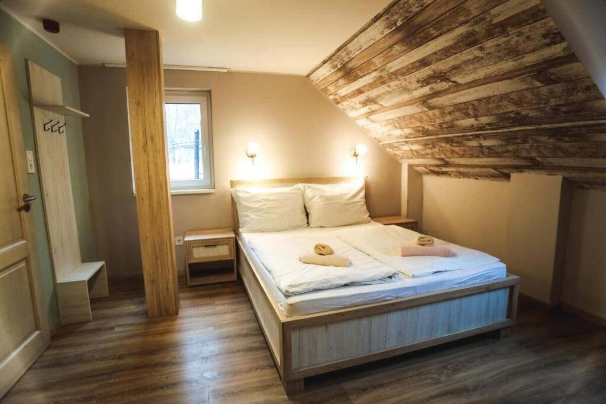 A bedroom with a wooden ceiling, a bed with white linens, a nightstand, and a window.