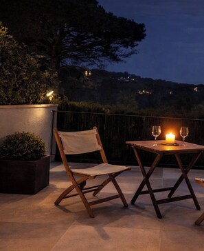A patio with a table and chair, a candle, and a view of a hill with lights. - Charming apartment with a pool in Calella de Palafrugell, on the Costa Brava (Calella de Palafrugell)