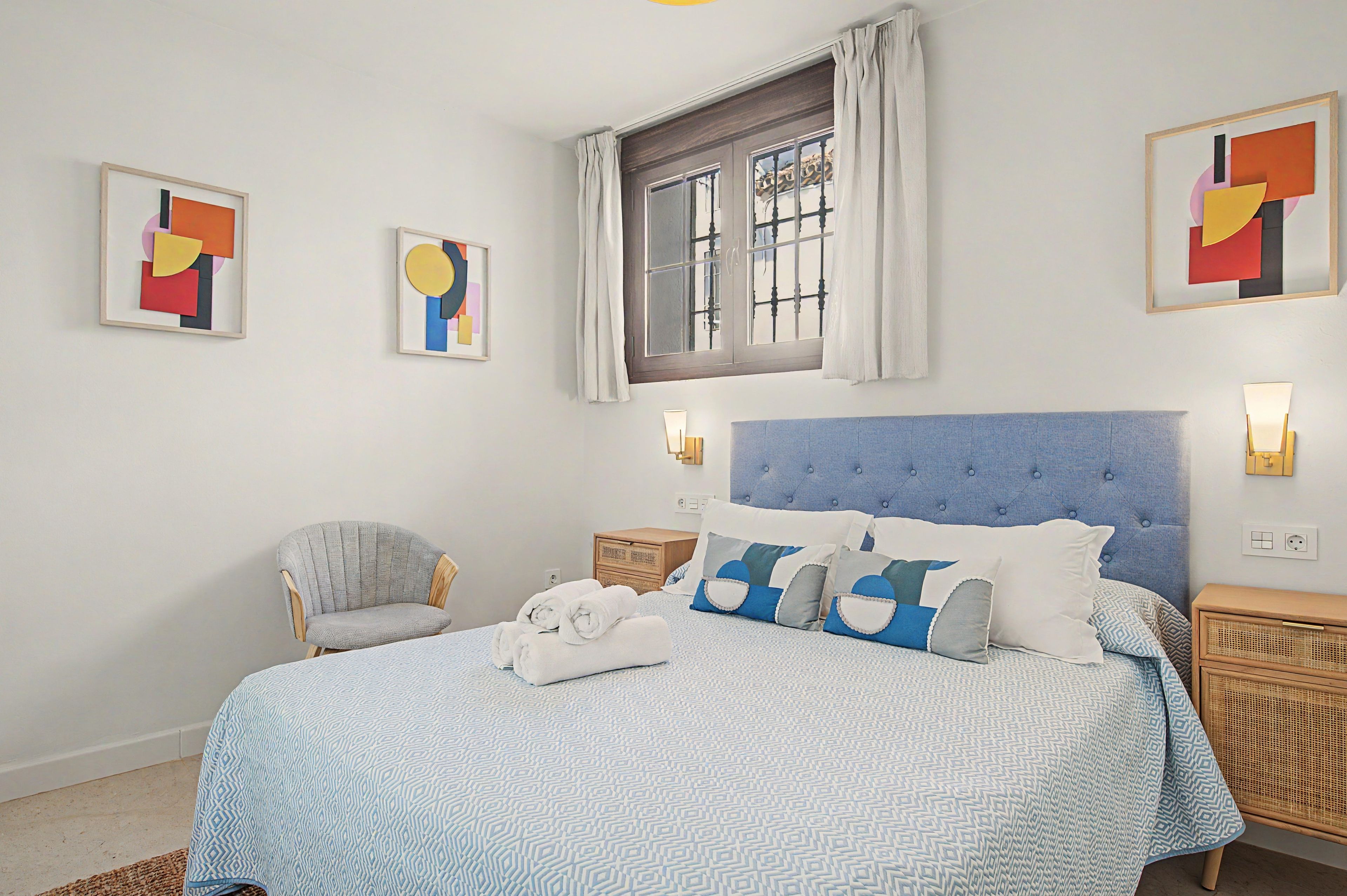 Un dormitorio con cabecera tapizada en azul, cama con ropa de cama estampada, una silla y obras de arte en las paredes.