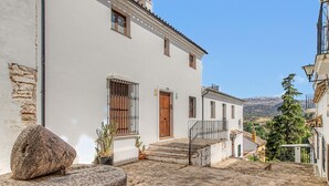 A white building with a wooden door and window shutters, a stone bench, and a view of a mountainous landscape.