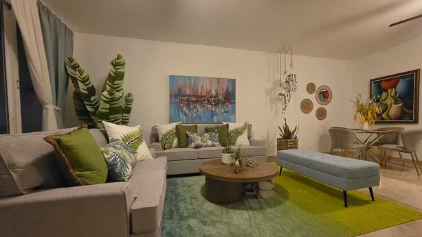 A modern living room with a sofa, a coffee table, and a blue ottoman. There is a painting on the wall and a dining area with chairs. - Punta Cana Oasis Apartment: Elegant and Peaceful (Punta Cana)