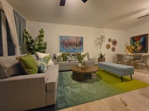 Ein modernes Wohnzimmer mit einer Couch, einem Couchtisch und einem blauen Hocker. An der Wand hängt ein Gemälde, und es gibt einen Essbereich mit Stühlen.