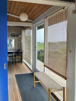 A room with a wooden floor, a bench, a desk with chairs, a window with blinds, and a door.