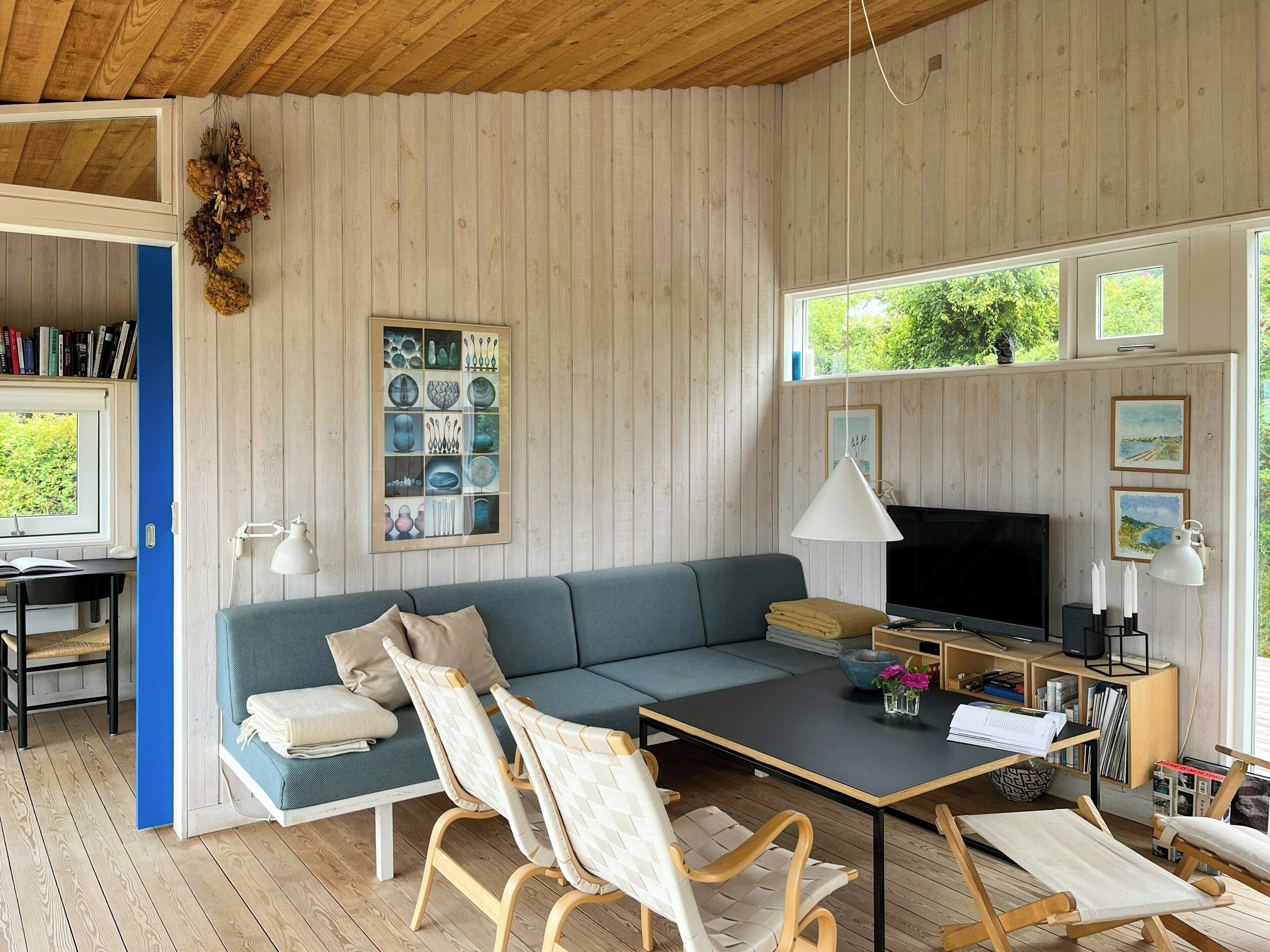 A cozy room with a wooden ceiling, a sofa, a TV, a desk with books, and a window with a view of greenery.