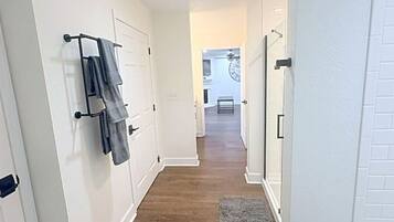 A hallway with white walls, wooden flooring, and a gray rug. There are doors on both sides, and a towel rack with towels is visible on the left wall.