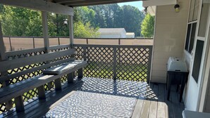 A covered outdoor deck with a wooden bench, a patterned rug, and a grill. - Lovely 2-bed 2-bath house with firepit in fenced in yard in charming Youngsville (Youngsville)