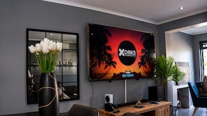 Un espace de bureau moderne doté d’un grand écran de télévision diffusant une image d’un coucher de soleil tropical, d’un bureau avec un ordinateur et d’un vase rempli de fleurs blanches.