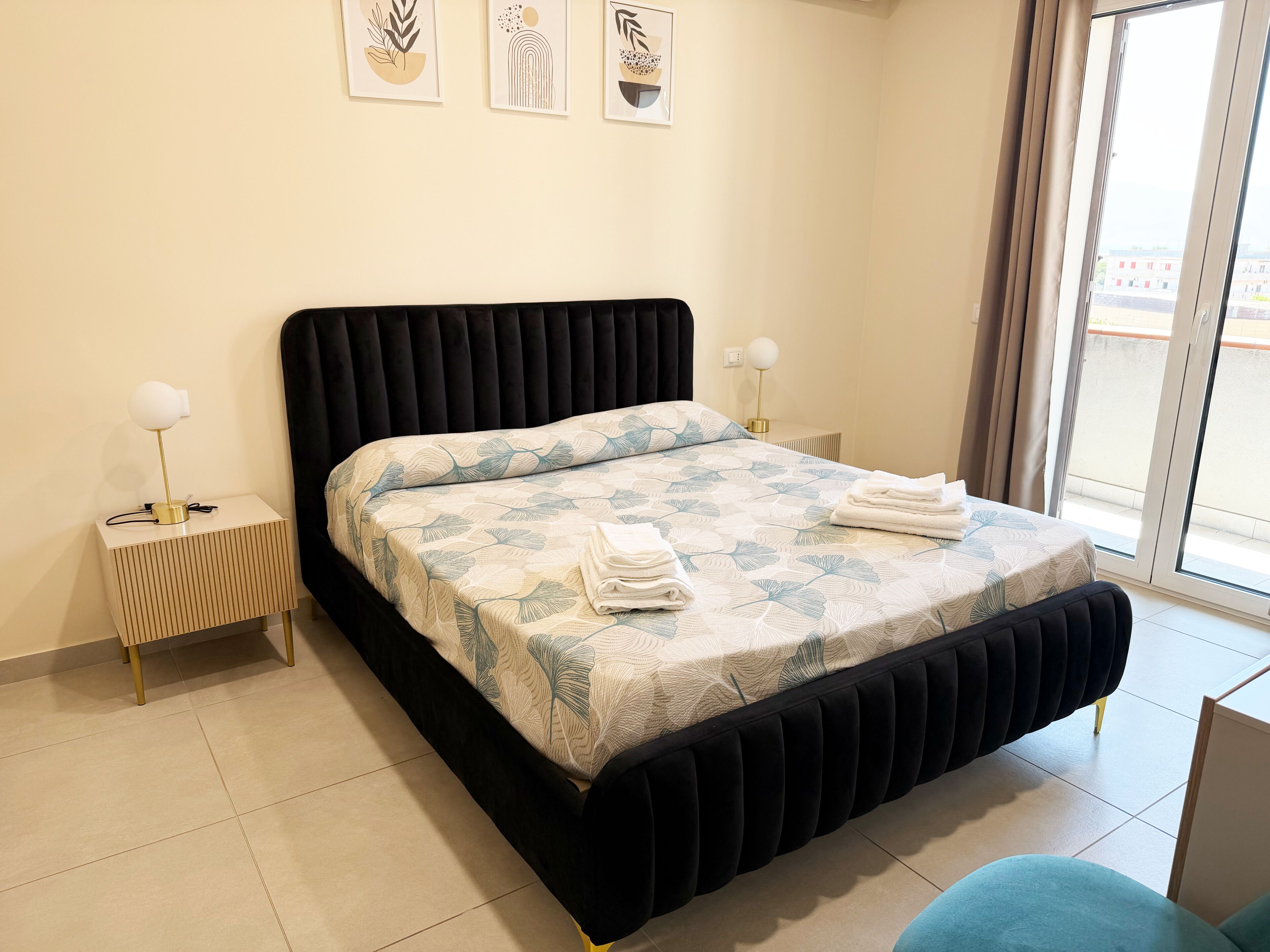 A bed with a patterned bedspread, two bedside tables with lamps, a window with curtains, and artwork on the wall.