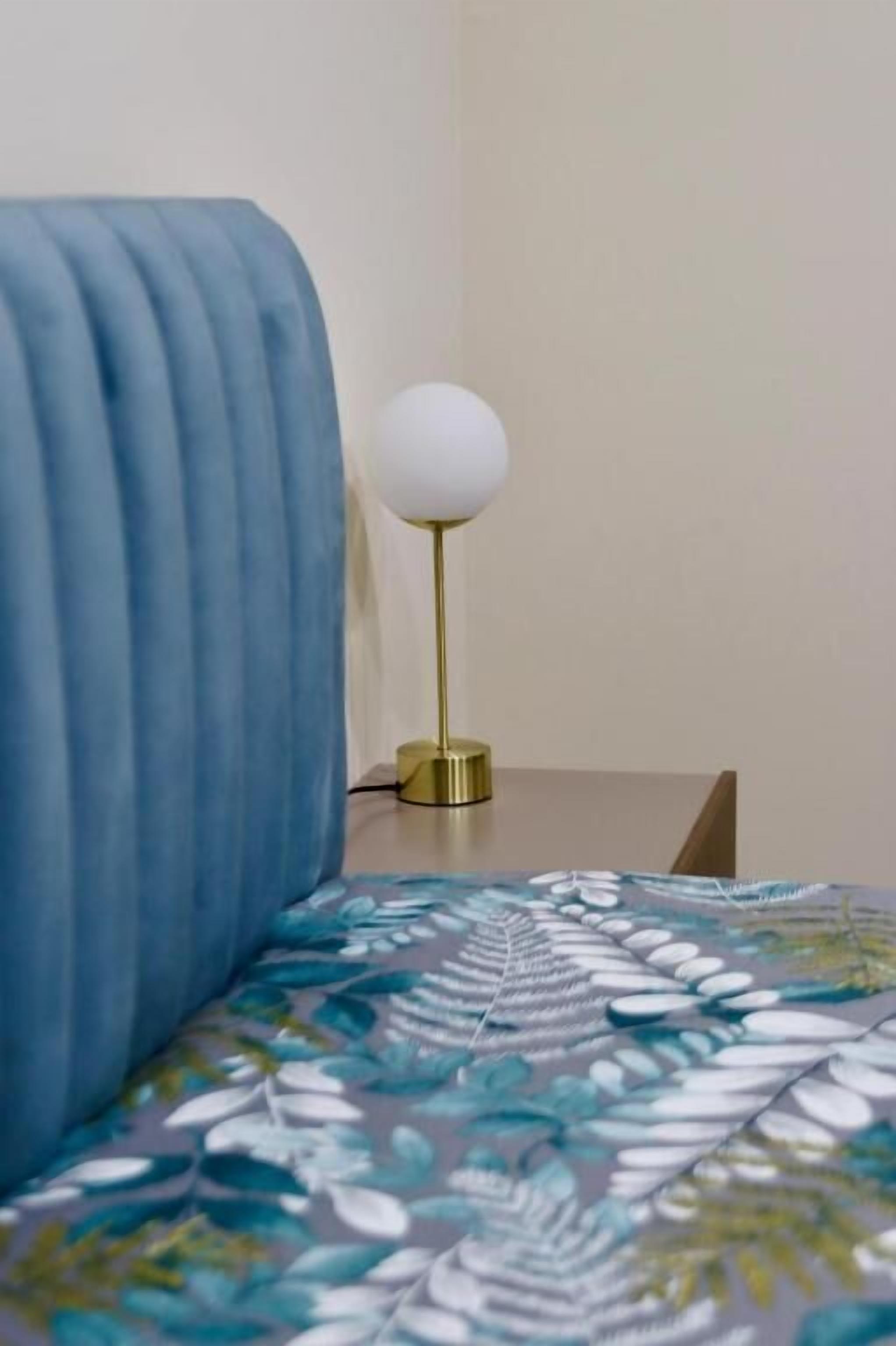 Ein Bett mit blauer Decke und einem blumigen Überwurf, eine Nachttischlampe mit weißem Schirm und ein Nachttisch aus Holz.
