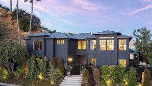 A modern two-story house with a dark exterior, multiple windows, and a well-maintained garden with various shrubs and a pathway leading to the entrance. - Sunset Manor - Designer Gated Estate Above Sunset Strip | Panoramic LA Views (Los Angeles)