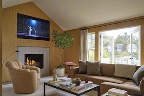 Ein Wohnzimmer mit Kamin, einer Couch, einem Couchtisch mit Büchern, einem an der Wand befestigten Fernseher und einem großen Fenster mit Blick.
