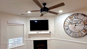 Ein Wohnzimmer mit Kamin, einem an der Wand befestigten Fernseher, einem Deckenventilator und einer großen Wanduhr.