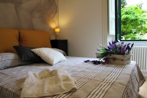 A neatly made bed with striped bedding, a bedside lamp, a nightstand, and a window with a view of greenery. - A relaxing green space in the city center (Salerno)