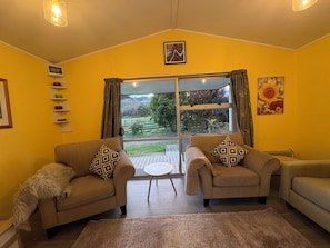 A living room with two armchairs, a sofa, a small round table, and a large window with curtains. - Cottage at Flat Hills Cafe and Tourist Park (self-contained) (Ōhingaiti)
