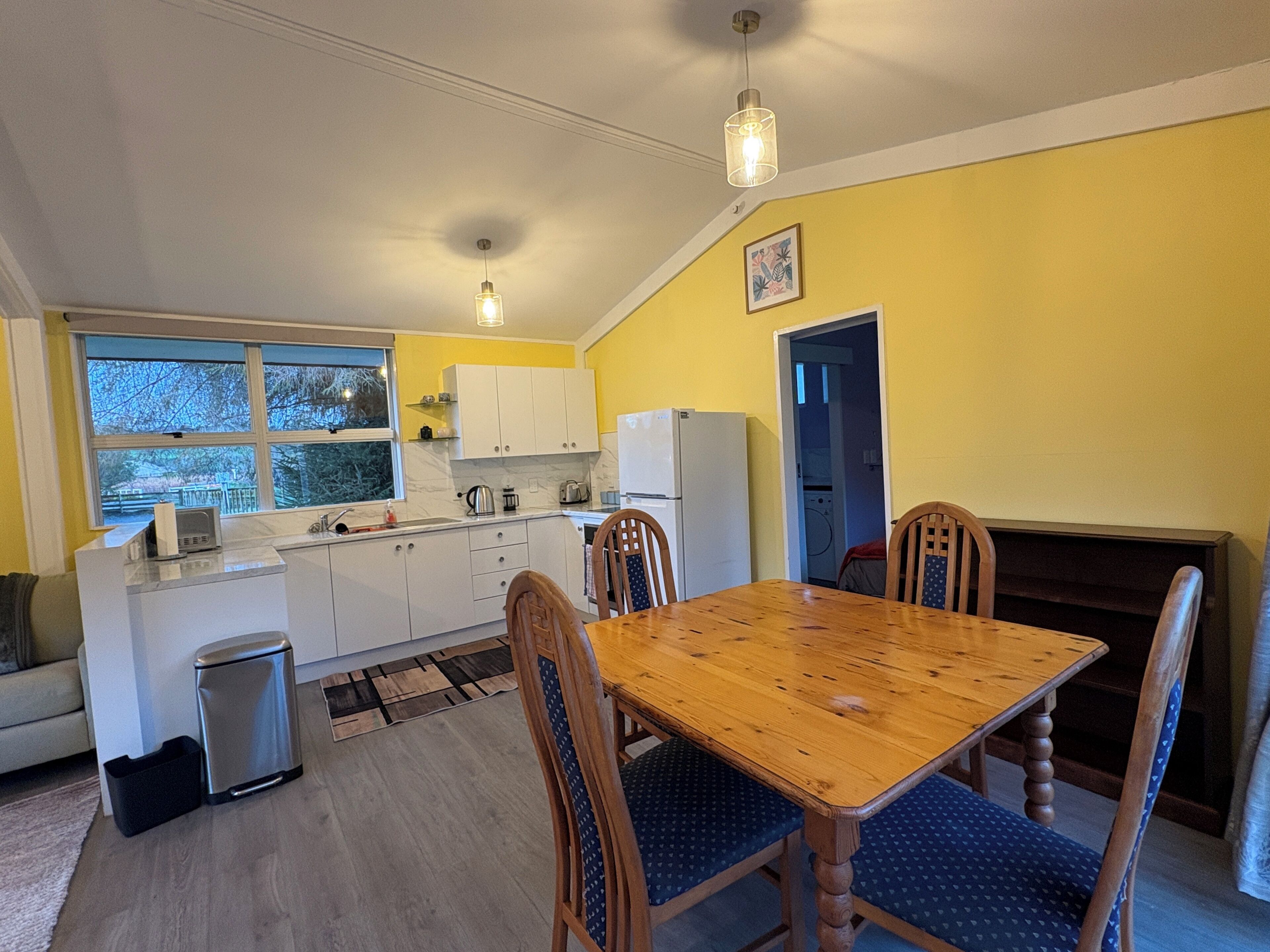 A compact kitchen with white cabinets, a dining table with blue upholstered chairs, and a window with a view of trees.