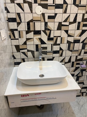 A bathroom with a white sink on a pedestal, a geometric patterned wall, and a visible light switch. - Happy Home Stay (Jammu)