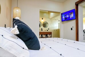 Une chambre à coucher avec un lit, un téléviseur fixé au mur, un luminaire suspendu et une salle de bain visible à travers une porte ouverte.