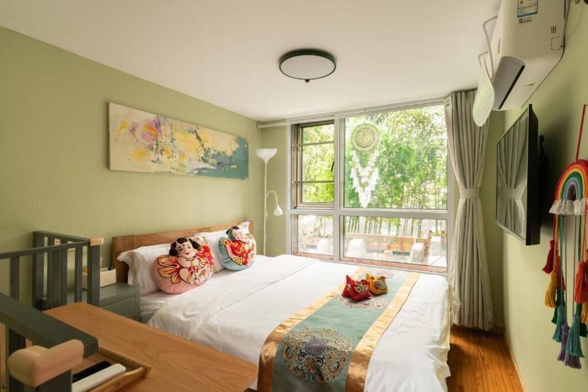 Un dormitorio con cama, escritorio, ventana con vistas a vegetación y un cuadro en la pared.
