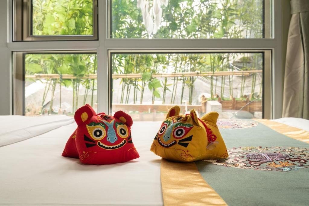 A bed with two decorative pillows, a patterned bedspread, and a view of greenery through large windows.