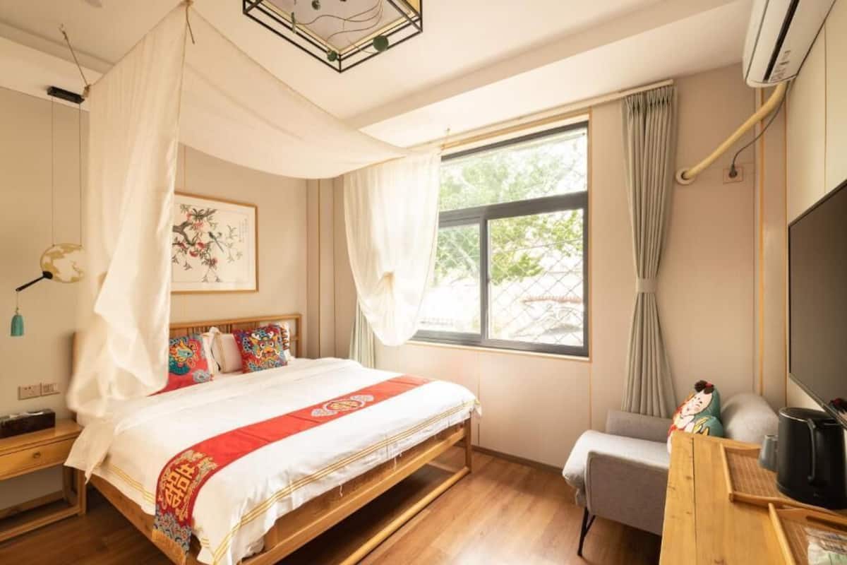 Un dormitorio con cama, mesita de noche, silla, ventana y una obra de arte.