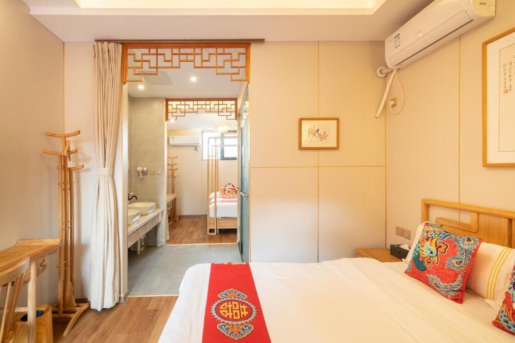 A hotel room with a bed, a wooden headboard, a bedside table, a wall-mounted air conditioner, and a bathroom visible through an open door.