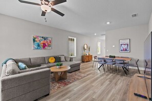 A modern living room with a grey sofa, a coffee table, and a dining area with chairs and a table. - Luxury Villa w/ Pool & Gym Near Disney (Kissimmee)