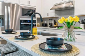 A modern kitchen with a stainless steel refrigerator, a built-in oven, and a sink. There are black bowls on placemats, and a vase with yellow flowers on the counter. - Luxury Villa w/ Pool & Gym Near Disney (Kissimmee)