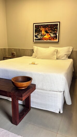 Una cama bien tendida con unacolcha blanca, un cuenco de madera sobre una mesa y un cuadro en la pared.