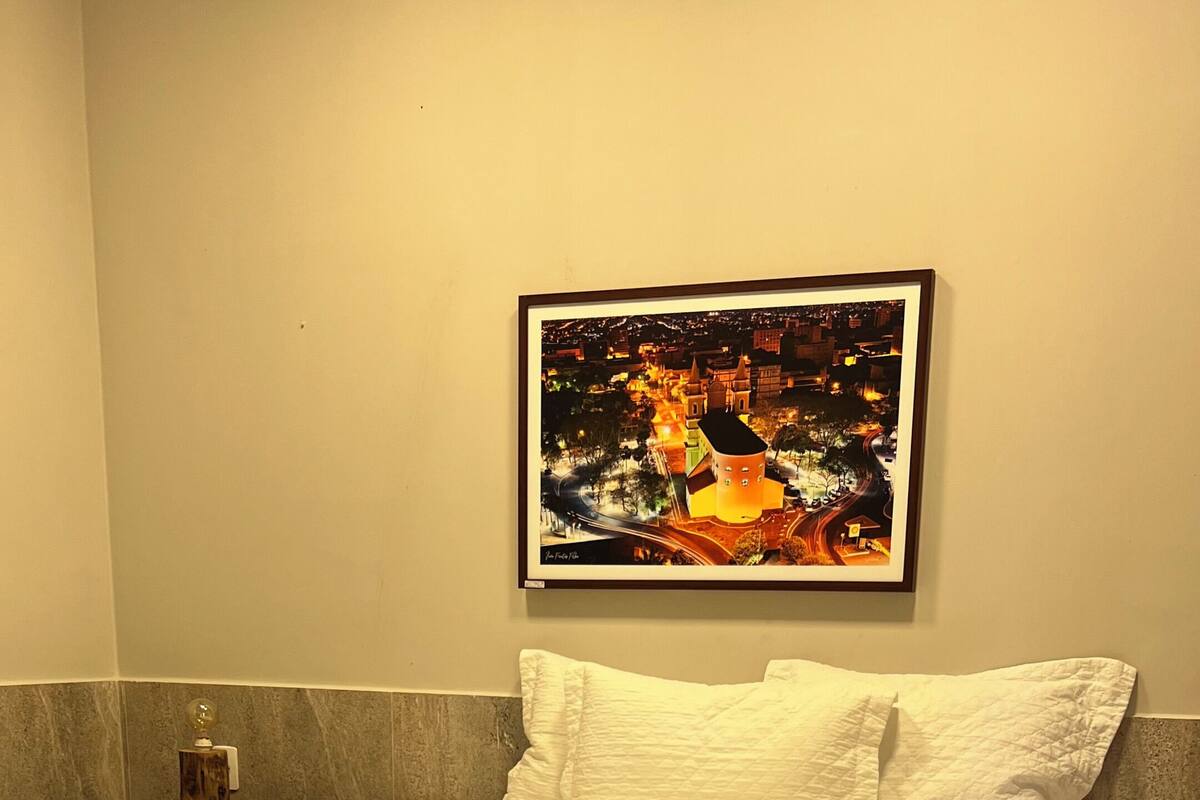 A neatly made bed with white bedding, a wooden bowl on a table, and a framed picture on the wall.