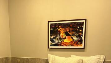 Uma cama bem arrumada com roupa de cama branca, uma tigela de madeira sobre uma mesa e um quadro pendurado na parede.
