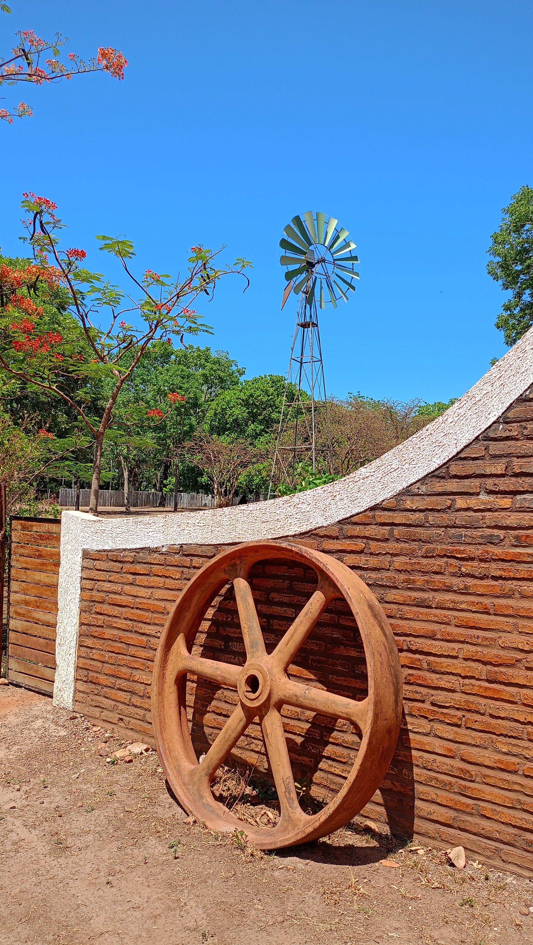 Uma grande roda de madeira, uma parede de tijolos e um moinho de vento ao fundo.