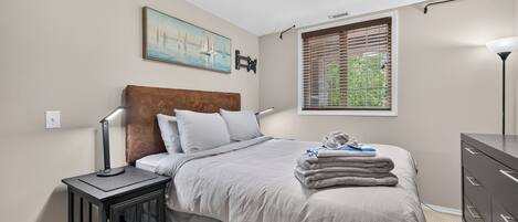 A bedroom with a bed, nightstand, dresser, window with blinds, and a painting on the wall.