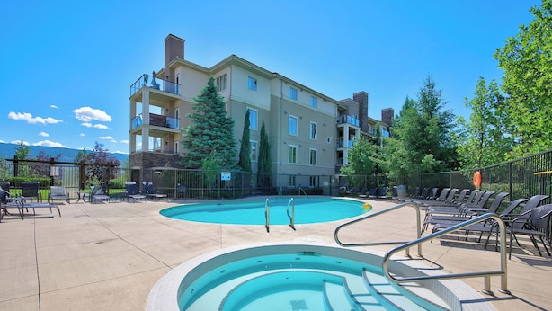 A swimming pool with a circular hot tub, surrounded by lounge chairs and a multi-story building with balconies. - Mountain View w Pool and Hot Tub 2213 (Kelowna)