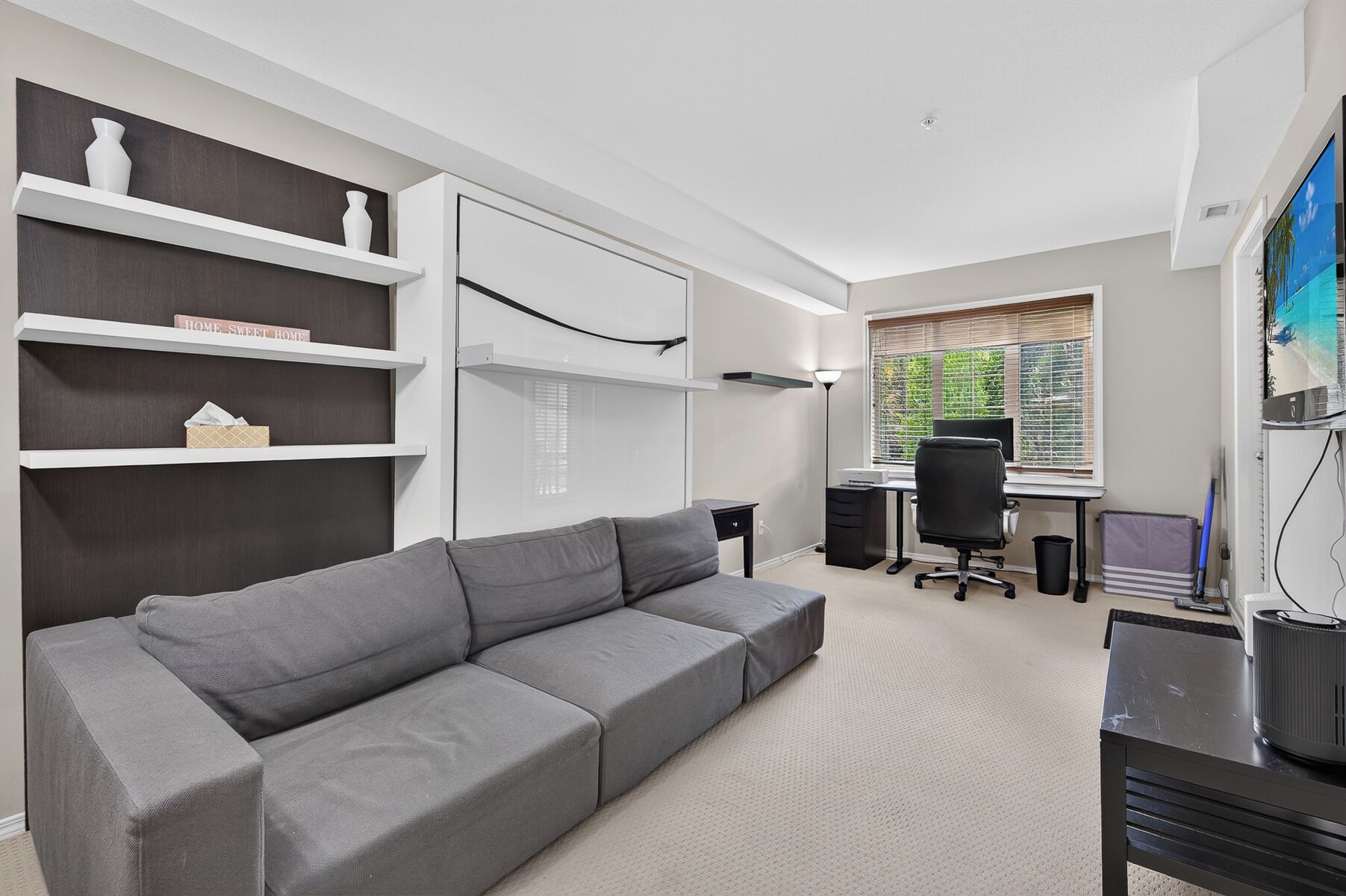 A modern room with a grey sofa, white shelving, a desk with a computer, and a window with blinds.