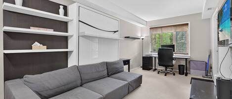 A modern room with a grey sofa, white shelving, a desk with a computer, and a window with blinds.