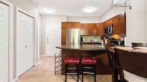 A kitchen with wooden cabinets, a stainless steel refrigerator, a microwave, and a bar area with stools. - Mountain View w Pool and Hot Tub 2213 (Kelowna)