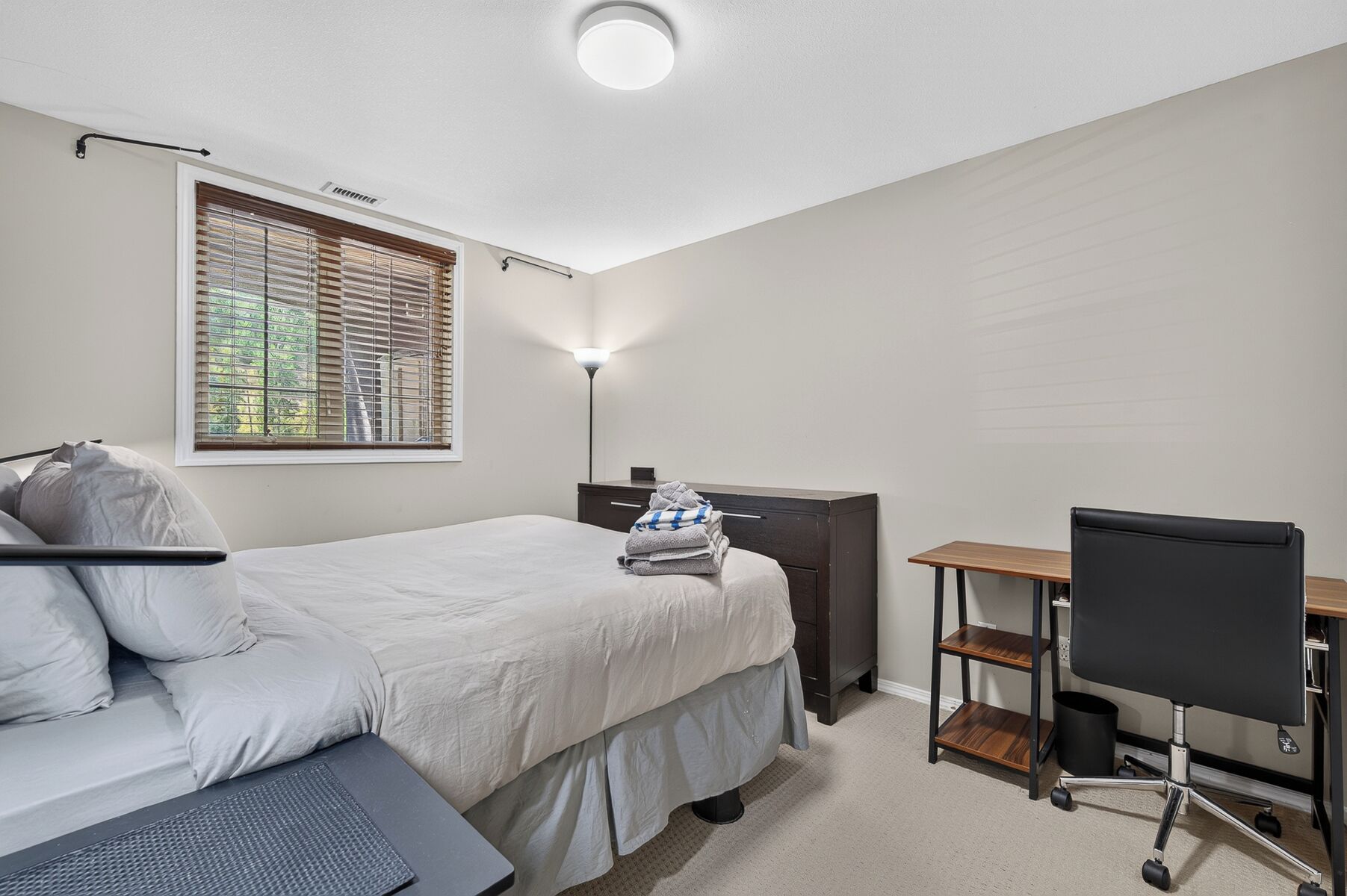 A bedroom with a bed, bedside table, desk, chair, and a window with blinds.