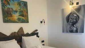 En pænt redt seng med hvide sengetøj og håndklæder, et billede af et tropisk landskab, og en vægmonteret lampe.