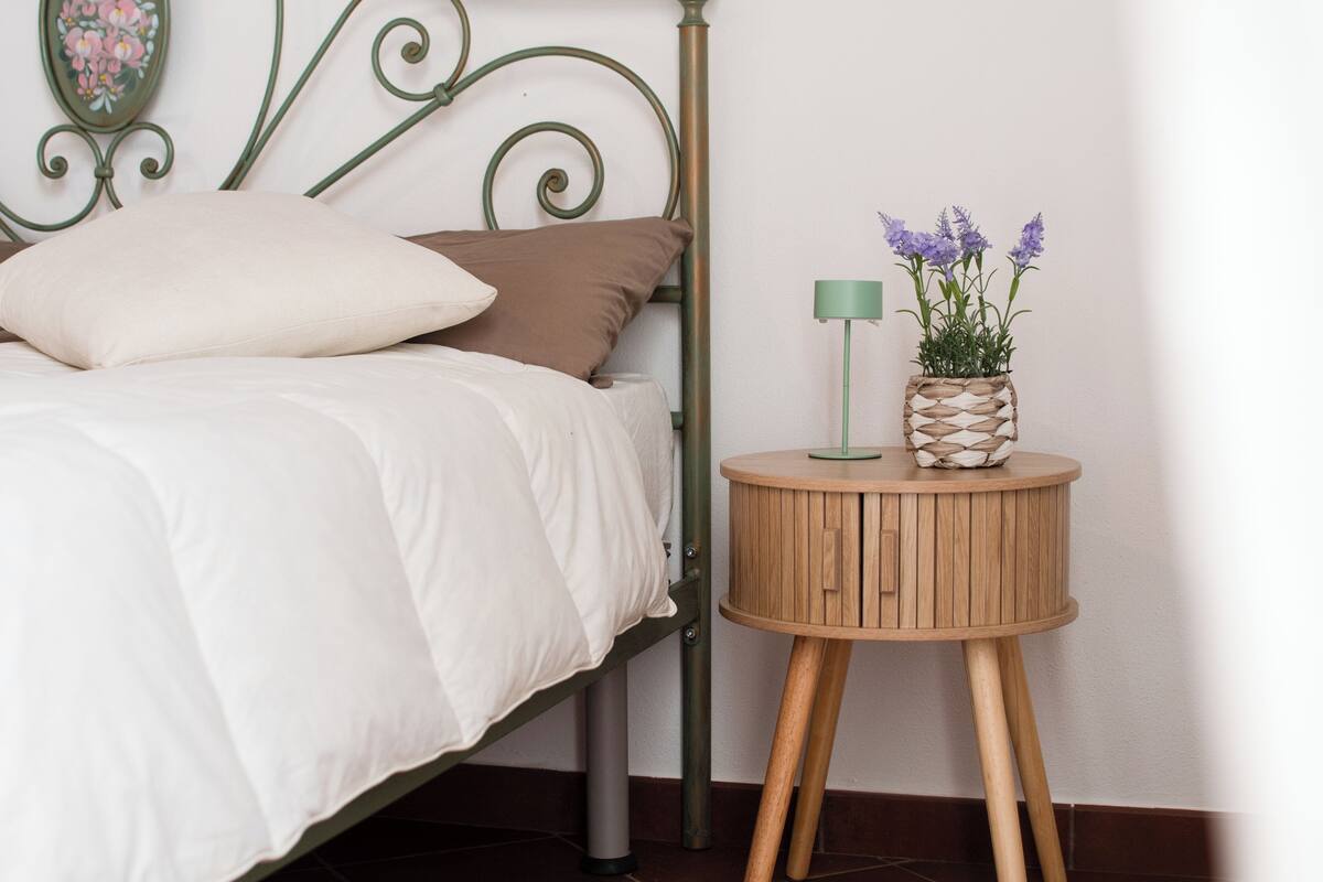A bed with pillows, a bedside table with a plant, and a wall-mounted decorative item.