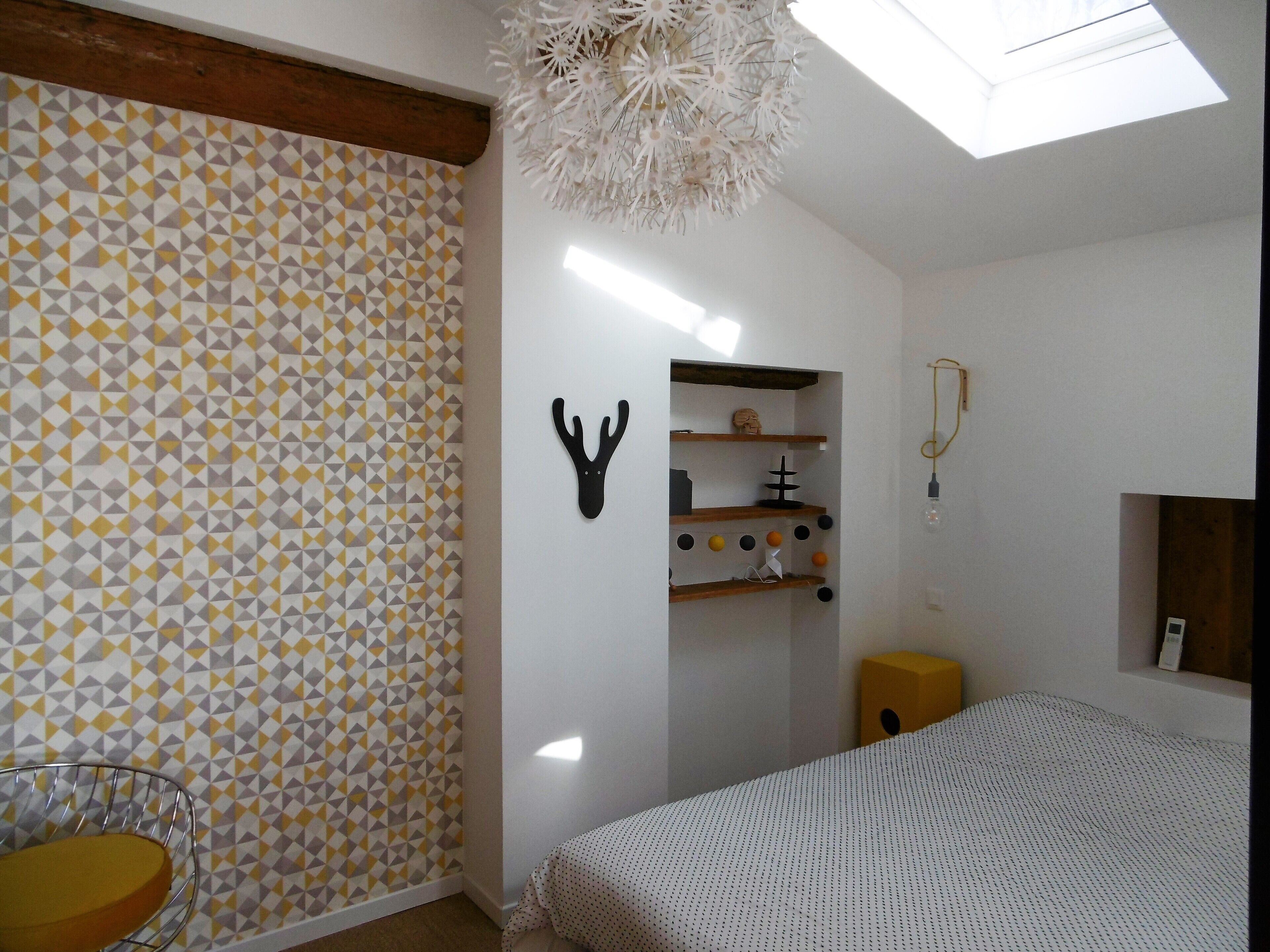 Une chambre avec un mur orné d’un motif géométrique, un lit, un luminaire au plafond et une lucarne.