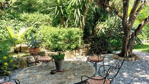 A patio area with a brick floor, surrounded by potted plants and trees, and furnished with metal chairs and a table. - Charming holiday cottage with pool-pétanque-table tennis-foosball-BBQ-parking (Roujan)