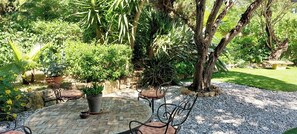 A patio area with a brick floor, surrounded by potted plants and trees, and furnished with metal chairs and a table. - Charming holiday cottage with pool-pétanque-table tennis-foosball-BBQ-parking (Roujan)