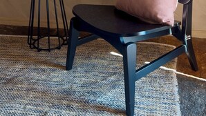 Ein Zimmer mit einer strukturierten, beige farbenen Wand, einem runden schwarzen Tisch mit einem weißen Gegenstand, einem schwarzen Stuhl mit rosa Kissen und einem gemusterten Teppich.
