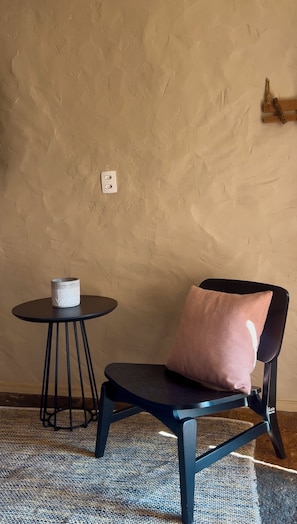 Huoneessa on struktuuripinnoitettu beige seinä, pyöreä musta pöytä, jonka päällä on valkoinen esine, musta tuoli, jossa on vaaleanpunainen koristetyyny, ja kuviollinen matto.
