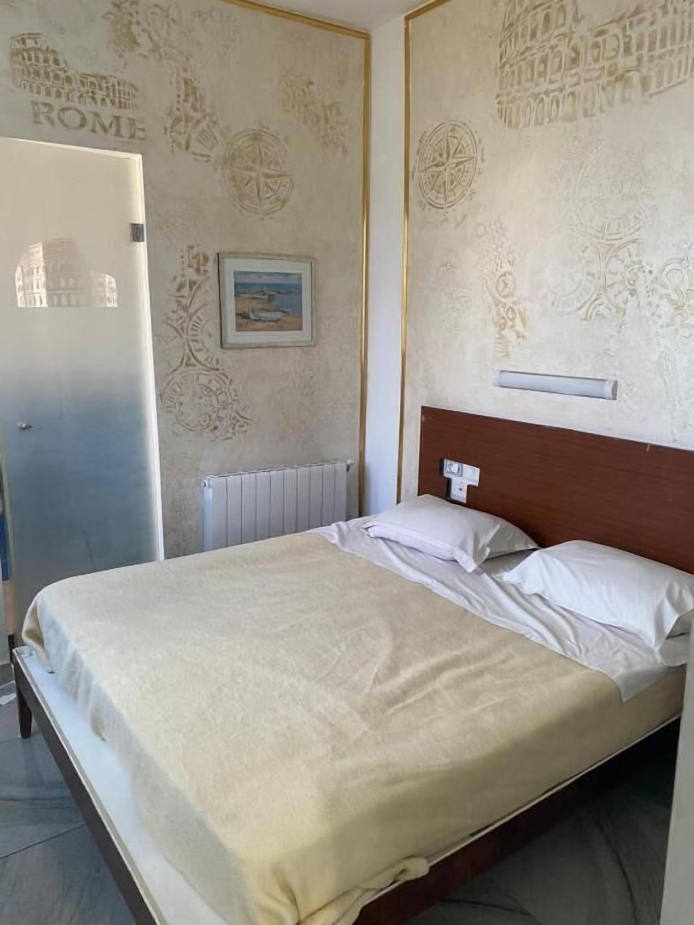 A bedroom with a bed, a nightstand, a wall-mounted light, and a door labeled 'ROME'.