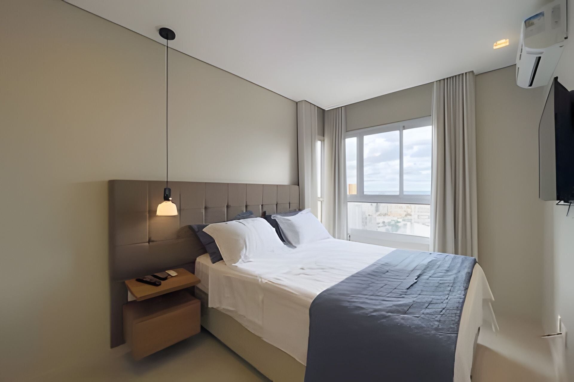 Quarto de hotel moderno com uma cama grande, mesa de cabeceira, luminária pendente, janela com cortinas e ar condicionado fixado na parede.