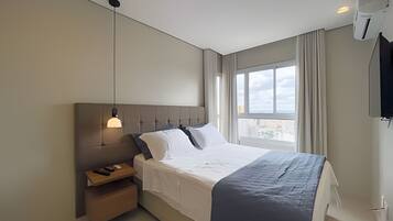 Quarto de hotel moderno com uma cama grande, mesa de cabeceira, luminária pendente, janela com cortinas e ar condicionado fixado na parede.